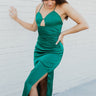 Full body side view of model wearing the Life Of The Party Maxi Dress which has an emerald green satin material, a deep open neckline, thin adjustable straps, a side drape detail with a slit, an open back, and a back zipper closure.