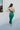 Full body back view of model wearing the Life Of The Party Maxi Dress which has an emerald green satin material, a deep open neckline, thin adjustable straps, a side drape detail with a slit, an open back, and a back zipper closure.
