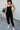 Full body front view of model wearing the Next Adventure Jumpsuit in Black, which features a black linen material, a square neck, a sleeveless design with tie straps, two front and back pockets, and elastic ankles. Worn over white tank.