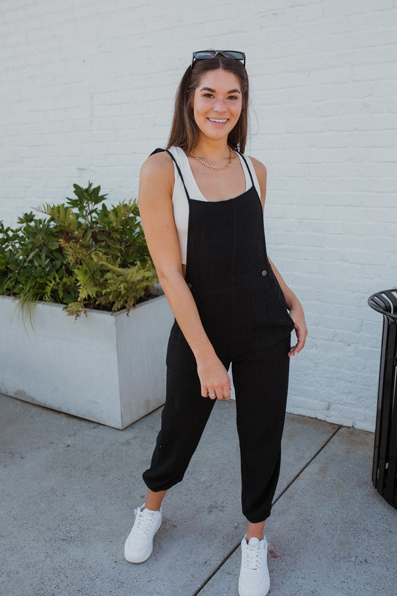 Full body front view of model wearing the Next Adventure Jumpsuit in Black, which features a black linen material, a square neck, a sleeveless design with tie straps, two front and back pockets, and elastic ankles. Worn over white tank.