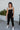 Full body front view of model wearing the Next Adventure Jumpsuit in Black, which features a black linen material, a square neck, a sleeveless design with tie straps, two front and back pockets, and elastic ankles. Worn over white tank.