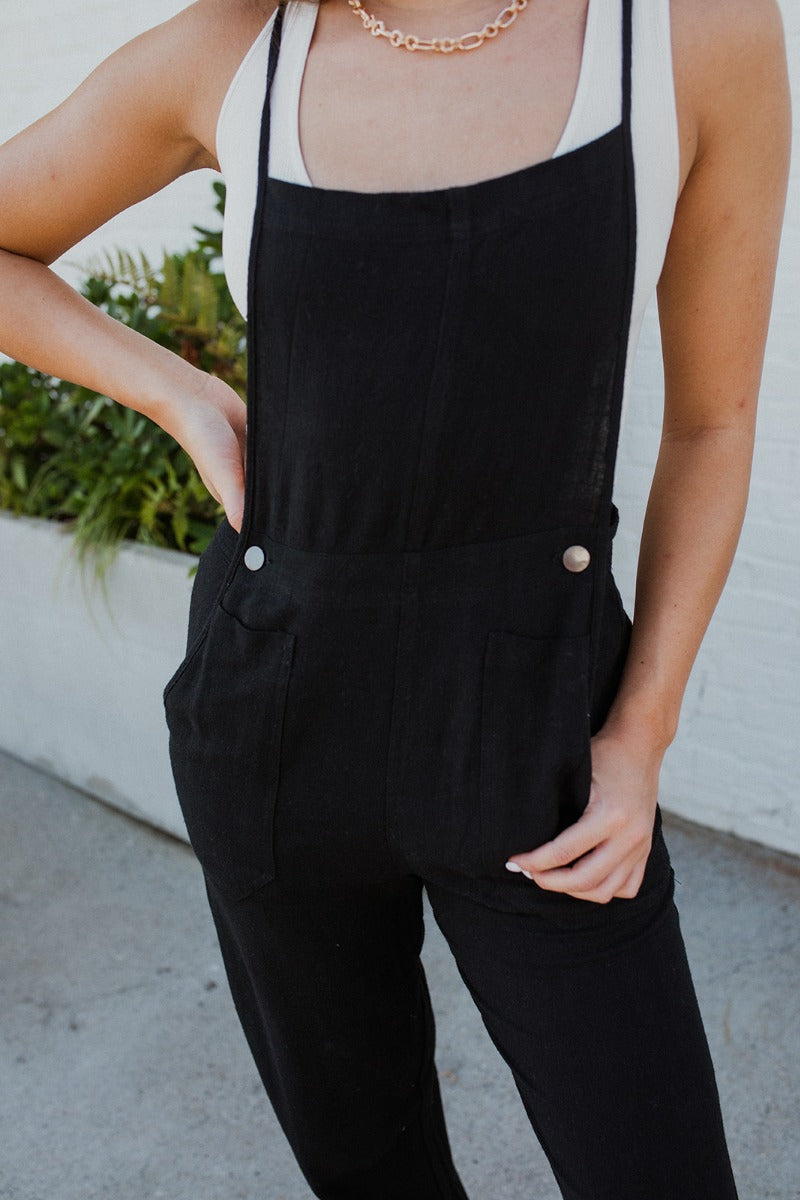 Close-up front view of model wearing the Next Adventure Jumpsuit in Black, which features a black linen material, a square neck, a sleeveless design with tie straps, two front and back pockets, and elastic ankles. Worn over white tank.