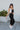 Full body side view of model wearing the Next Adventure Jumpsuit in Black, which features a black linen material, a square neck, a sleeveless design with tie straps, two front and back pockets, and elastic ankles. Worn over white tank.