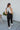 Full body back view of model wearing the Next Adventure Jumpsuit in Black, which features a black linen material, a square neck, a sleeveless design with tie straps, two front and back pockets, and elastic ankles. Worn over white tank.