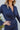 Close-up neckline view of model wearing the First Impressions Dress, which has a navy colored material, a collar neck, a surplice neckline, a long cuffed sleeve, a side draping detail, and a mini length.