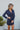 Front view of model wearing the First Impressions Dress, which has a navy colored material, a collar neck, a surplice neckline, a long cuffed sleeve, a side draping detail, and a mini length.
