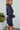 Side view of model wearing the First Impressions Dress, which has a navy colored material, a collar neck, a surplice neckline, a long cuffed sleeve, a side draping detail, and a mini length.