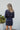 Back view of model wearing the First Impressions Dress, which has a navy colored material, a collar neck, a surplice neckline, a long cuffed sleeve, a side draping detail, and a mini length.