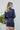 Back view of model wearing the First Impressions Dress, which has a navy colored material, a collar neck, a surplice neckline, a long cuffed sleeve, a side draping detail, and a mini length.