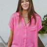 Front view of model wearing the First Class Top, which has a bubble gum pink material, a collar neckline, a button-up front, a front pocket, short cuffed sleeves, and a flowy fit