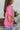 Side view of model wearing the First Class Top, which has a bubble gum pink material, a collar neckline, a button-up front, a front pocket, short cuffed sleeves, and a flowy fit