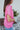 Side view of model wearing the First Class Top, which has a bubble gum pink material, a collar neckline, a button-up front, a front pocket, short cuffed sleeves, and a flowy fit