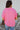 Back view of model wearing the First Class Top, which has a bubble gum pink material, a collar neckline, a button-up front, a front pocket, short cuffed sleeves, and a flowy fit
