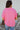 Back view of model wearing the First Class Top, which has a bubble gum pink material, a collar neckline, a button-up front, a front pocket, short cuffed sleeves, and a flowy fit