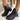 Side view of model's ankles- model is wearing the Harper Booties, that have black faux-leather uppers, v cut-outs on each side, a back zipper closure, lug soles, and a 2" heel. 
