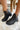 Side view of model's ankles- model is wearing the Harper Booties, that have black faux-leather uppers, v cut-outs on each side, a back zipper closure, lug soles, and a 2" heel. 