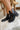 Side view of model's ankles- model is wearing the Harper Booties, that have black faux-leather uppers, v cut-outs on each side, a back zipper closure, lug soles, and a 2" heel. 