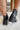 Back view of model's ankles- model is wearing the Harper Booties, that have black faux-leather uppers, v cut-outs on each side, a back zipper closure, lug soles, and a 2" heel. 