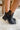 Side view of model's ankles- model is wearing the Harper Booties, that have black faux-leather uppers, v cut-outs on each side, a back zipper closure, lug soles, and a 2" heel. 
