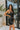 Side view of the City Walk Dress that features a black silky material, a cowl neckline, a sleeveless design with thin adjustable straps, a tie at the waist, a draped design, a back zipper closure, and a mini length.