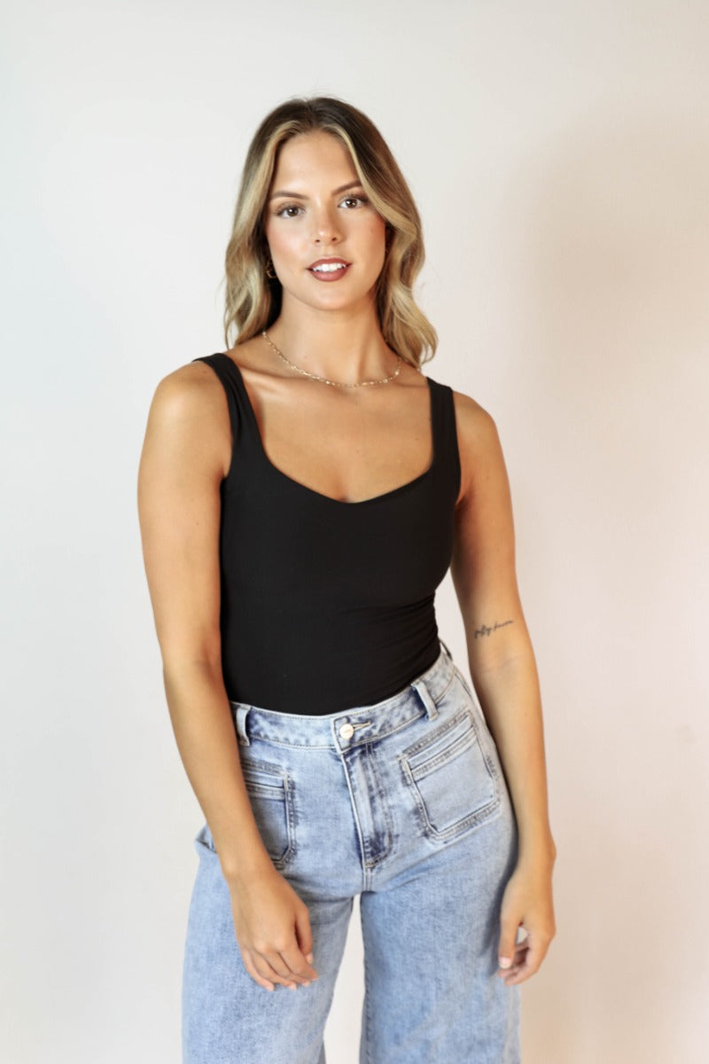 front view of model wearing the Avery Black Sleeveless Bodysuit that has black knit fabric, a scooped neckline, thick straps, and a thong bottom with snap closures.