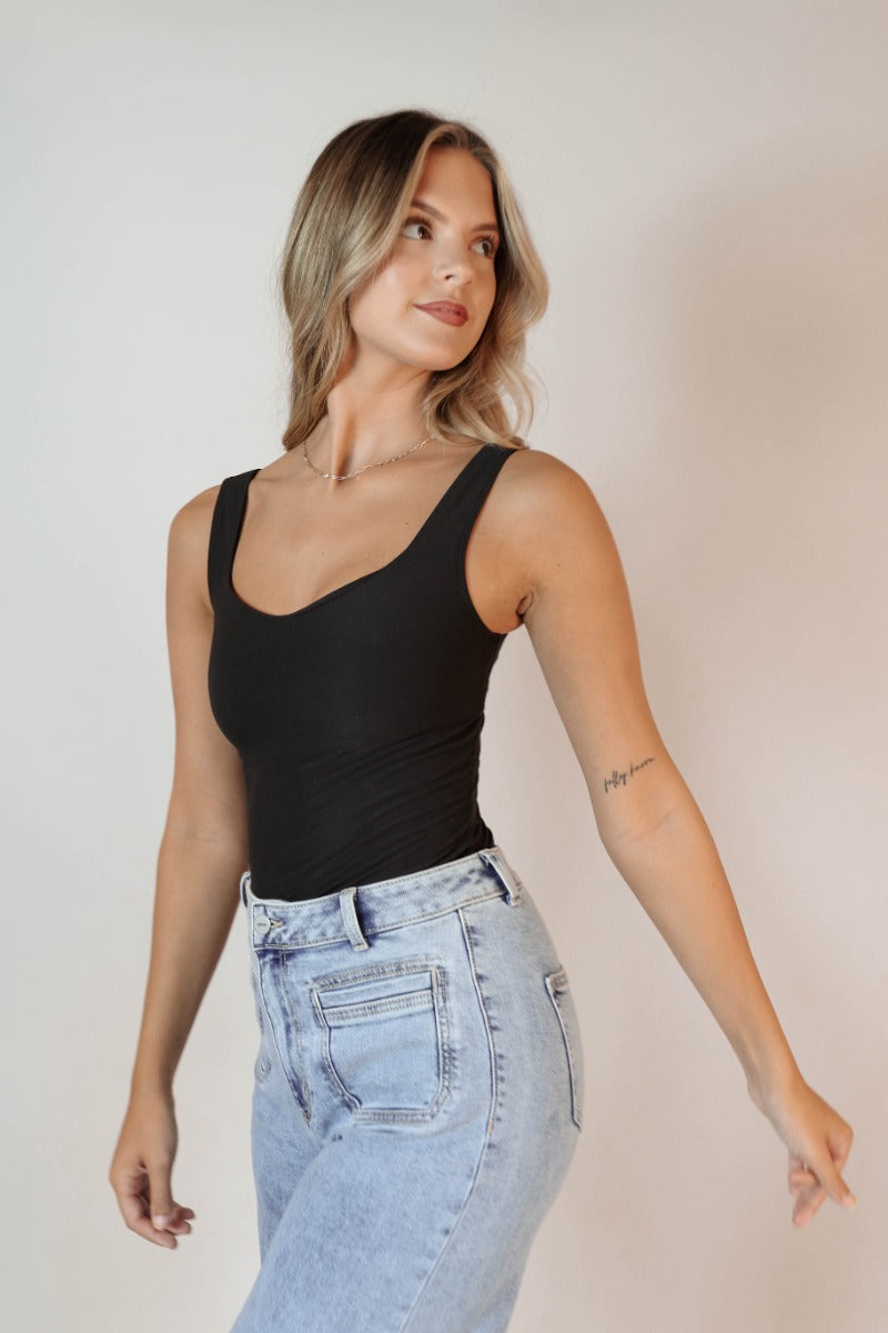 Side view of model wearing the Avery Black Sleeveless Bodysuit that has black knit fabric, a scooped neckline, thick straps, and a thong bottom with snap closures.
