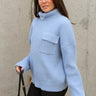 Side view of model wearing the Grace Light Blue Long Sleeve Sweater which features light blue cable knit fabric, ribbed hem, a ribbed turtleneck neckline, a left front chest folded pocket, dropped shoulders, and long sleeves.