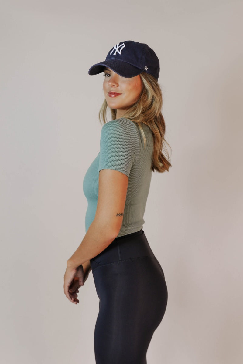 Side view of model wearing the Shae Sage Green One-Size Crop Top that has muted sage ribbed fabric, cropped waist, round neckline and short sleeves.