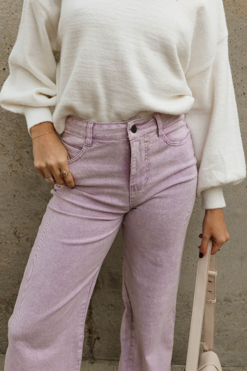 Close up view of model wearing the Emily Lavender Wide Leg Pants which features acid lavender denim-like fabric, two front pockets, a front zipper with a button closure, belt loops, two back pockets, and wide legs.