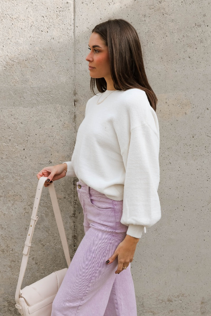 Side view of model wearing the Ella Cream Drawstring Cinch Long Sleeve Sweater which features white knit fabric, a round neckline, dropped shoulders, long balloon sleeves with ribbed cuffs, and a drawstring cinch detail in the back.