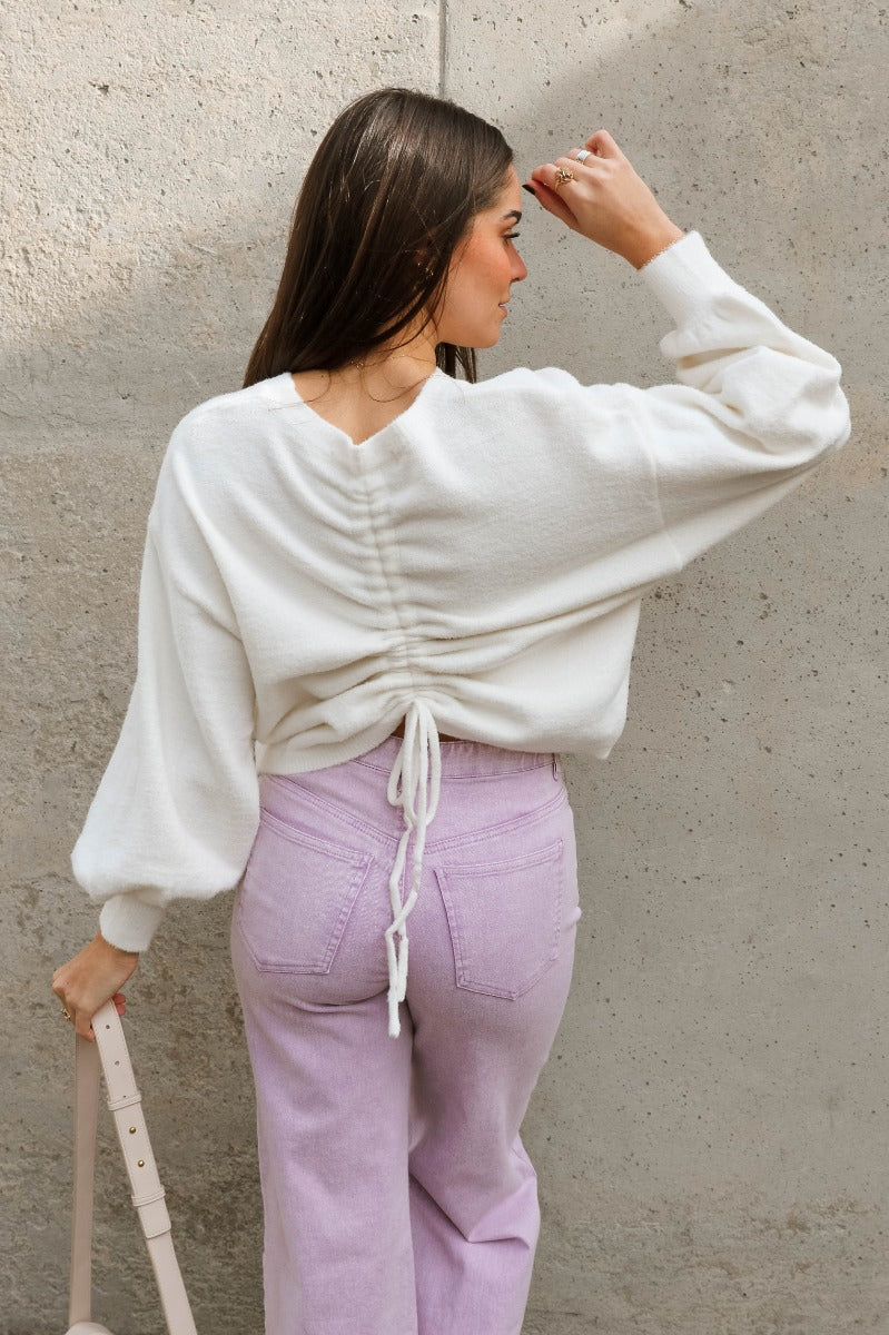 Back view of model wearing the Ella Cream Drawstring Cinch Long Sleeve Sweater which features white knit fabric, a round neckline, dropped shoulders, long balloon sleeves with ribbed cuffs, and a drawstring cinch detail in the back.
