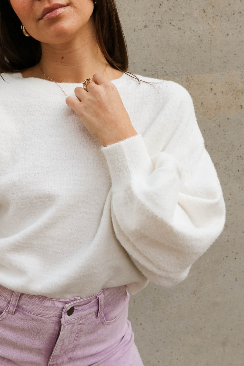 Close up view of model wearing the Ella Cream Drawstring Cinch Long Sleeve Sweater which features white knit fabric, a round neckline, dropped shoulders, long balloon sleeves with ribbed cuffs, and a drawstring cinch detail in the back.