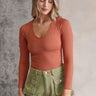 front view of model wearing the Kallie Rust Long Sleeve One-Size Bodysuit that has rust orange ribbed fabric, a v-neckline, long sleeves, and a thong bottom with a snap closure.