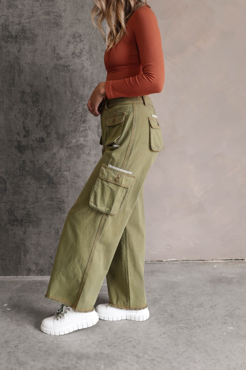 Side view of model wearing the Kimberly Olive Wide Leg Cargo Pants that have olive green denim-like fabric, brown contrast stitching, distressing, cargo pockets, a front zipper, belt loops, and wide legs.