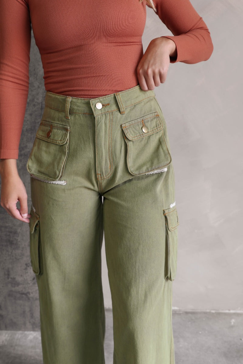 Close upper front view of model wearing the Kimberly Olive Wide Leg Cargo Pants that have olive green denim-like fabric, brown contrast stitching, distressing, cargo pockets, a front zipper, belt loops, and wide legs.