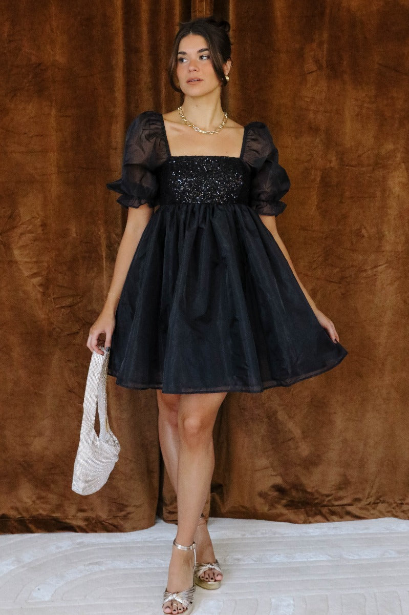 Full body front view of model wearing the Ariah Black Sequin Puff Sleeve Mini Dress that has black sheer fabric with a black sequin upper, babydoll style, a square neck, and puff half sleeves.