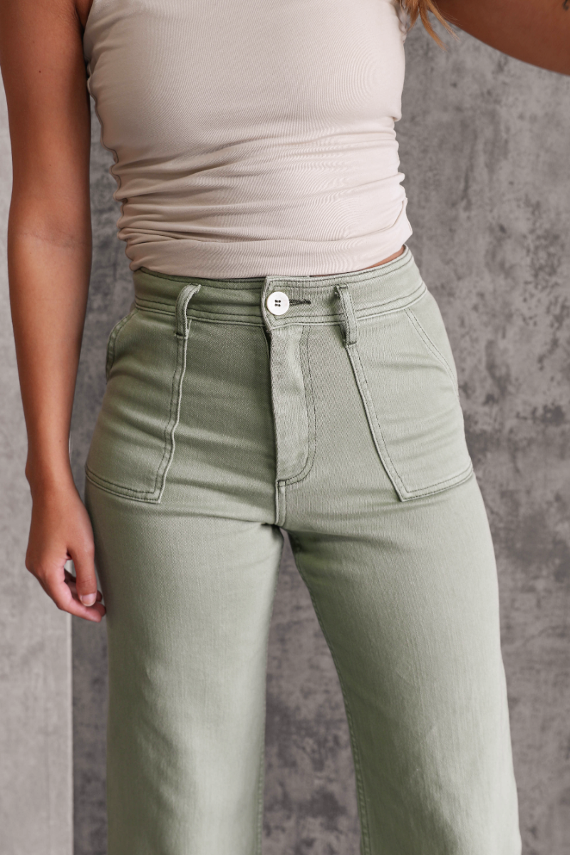 Close up view of model wearing the Alexa Sage High-Waisted Wide Leg Pants which features sage denim fabric, two front pockets, two back pockets, front zipper with button closure, belt loops and wide pant legs.