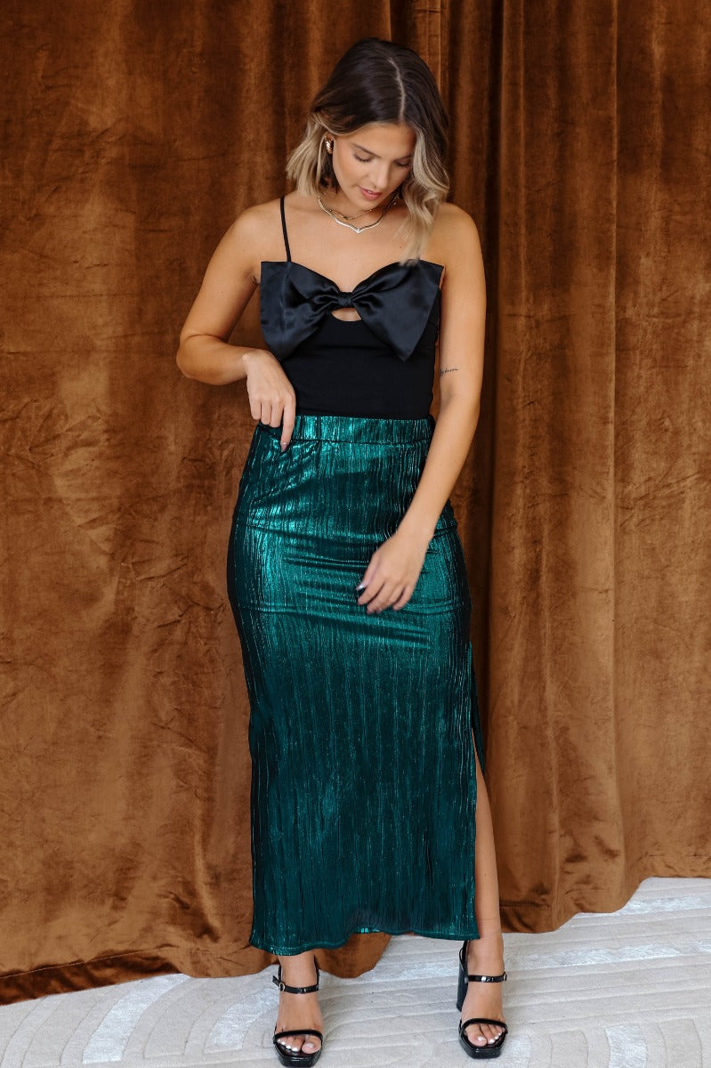Full body view of model wearing the Lainey Green Slit Midi Skirt which features teal and black shimmer pleated fabric, midi length, slit on the side, thigh length fuchsia lining and elastic waistband.