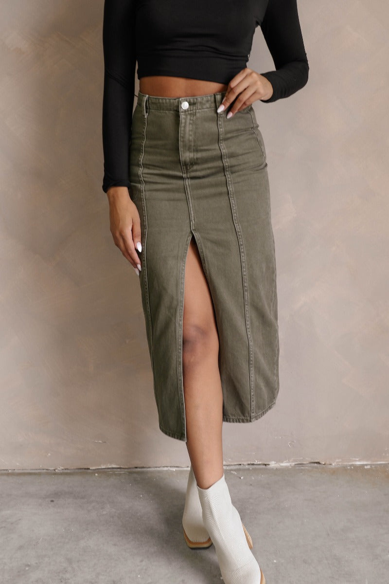 front view of model wearing the Kara Olive Denim Front Slit Midi Skirt that has olive green denim fabric, midi length, a front slit, a front zipper with a button closure, and belt loops.