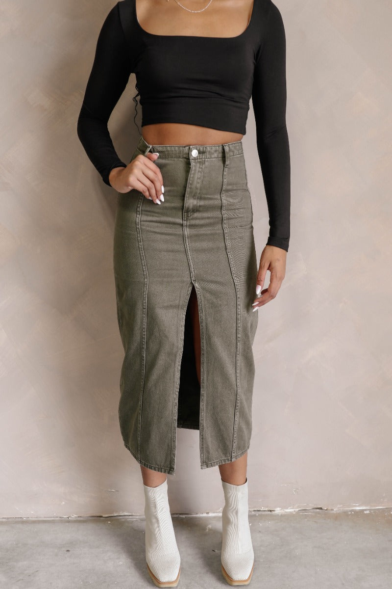 front view of model wearing the Kara Olive Denim Front Slit Midi Skirt that has olive green denim fabric, midi length, a front slit, a front zipper with a button closure, and belt loops.