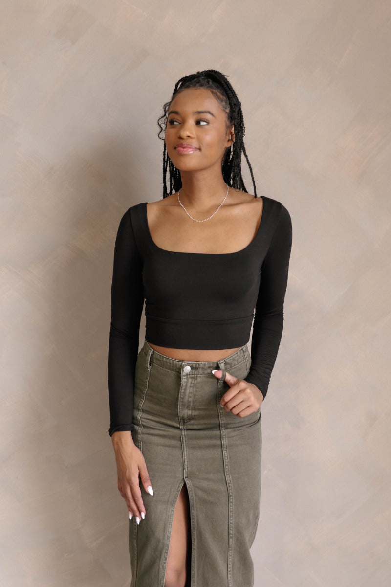 front view of model wearing the Greta Black Basic Long Sleeve Crop Top that has black knit fabric, a cropped waist, a thick band hem, a square neckline, and long sleeves.