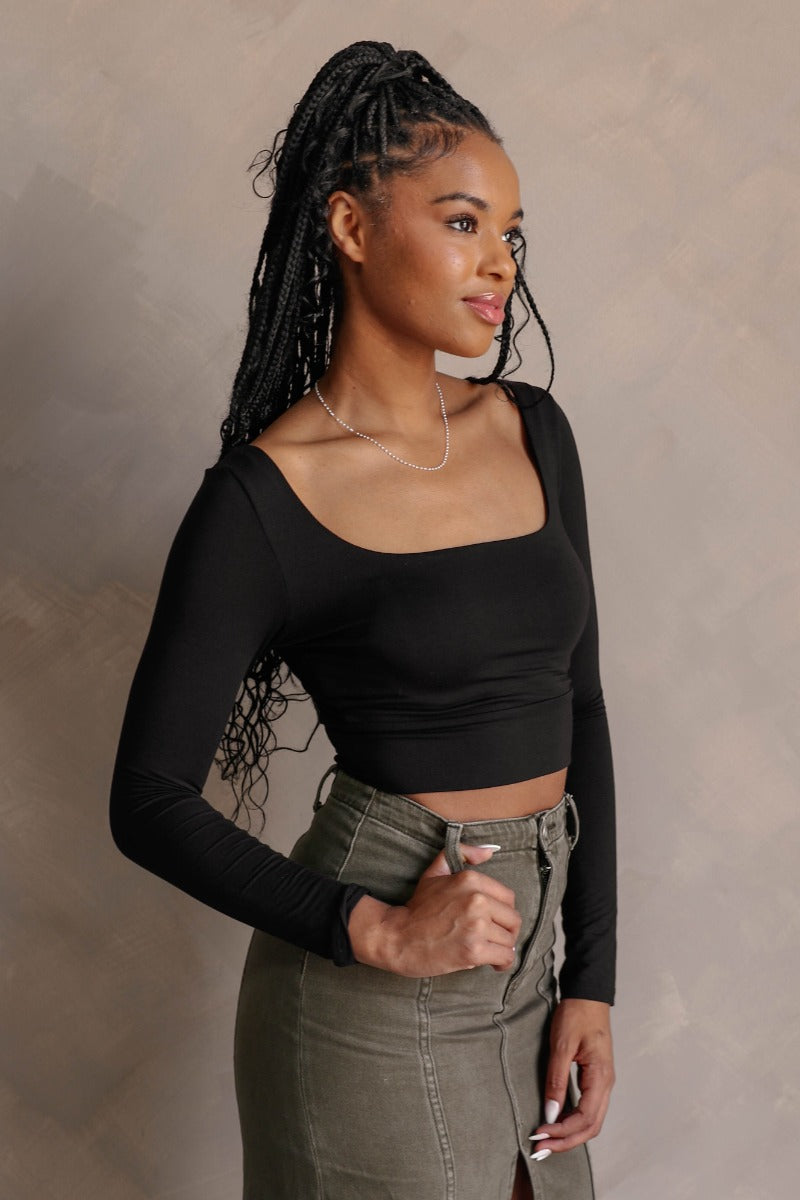 Side view of model wearing the Greta Black Basic Long Sleeve Crop Top that has black knit fabric, a cropped waist, a thick band hem, a square neckline, and long sleeves.
