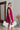 Side view of the Perfect Pair Dress that features a magenta material, a high rounded neckline with a ruffle detail, a short ruffle sleeve, a pleated top, a tiered design, a back button closure, a flowy fit, and a mini length.