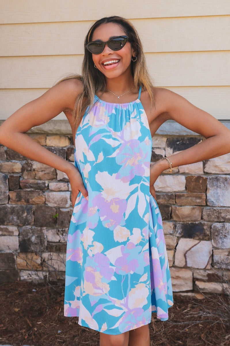 Front view of model wearing the Bahama Blues Floral Dress that has blue fabric with a pink, light blue, light pink and tan floral pattern, mini length, a halter neck, a sleeveless design, and a back keyhole with a tie closure.