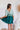 Back view of the Sweet Summer Romper that features a multi-colored design, a V neckline, a half cuffed sleeve, a three tiered design, two side pockets, a shorts lining, a V back with a tie detail, a flowy fit, and a mini length.