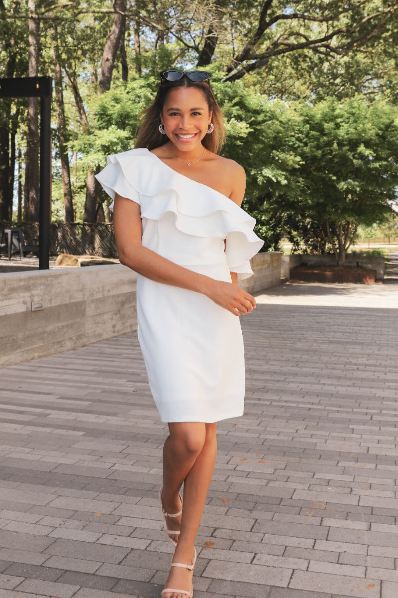 Front view of model wearing the Hopeless Romantic Dress that features white fabric, mini length, white lining, an off-the-shoulder ruffle design, and a monochromatic back zipper with a hook closure.