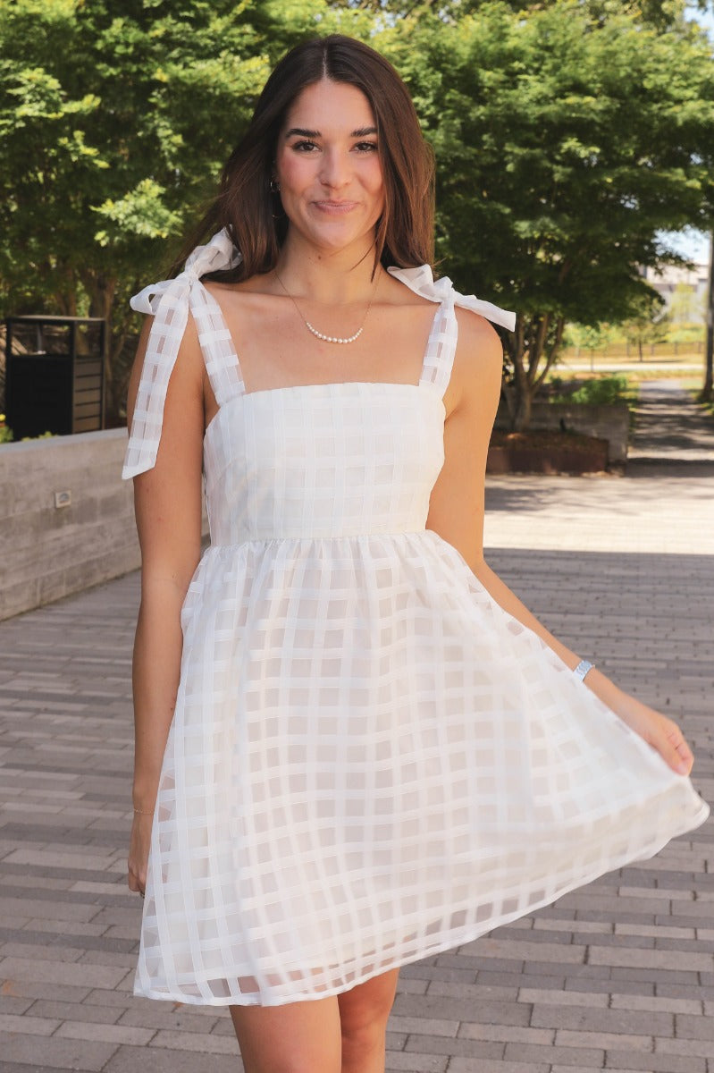 Front view of model wearing the Time Of My Life Dress that has  off white sheer fabric, a monochromatic plaid design, mini length, a square neck, tie sheer straps, and a smocked back.