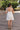 Full back view of model wearing the Time Of My Life Dress that has  off white sheer fabric, a monochromatic plaid design, mini length, a square neck, tie sheer straps, and a smocked back.