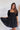 Frontal view of the Balanced Out Dress that features a black cotton material, a squared neckline, a half sleeve, a tiered babydoll design, a back button closure, a flowy fit, and a mini length.