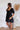 
Side view of the Balanced Out Dress that features a black cotton material, a squared neckline, a half sleeve, a tiered babydoll design, a back button closure, a flowy fit, and a mini length.
